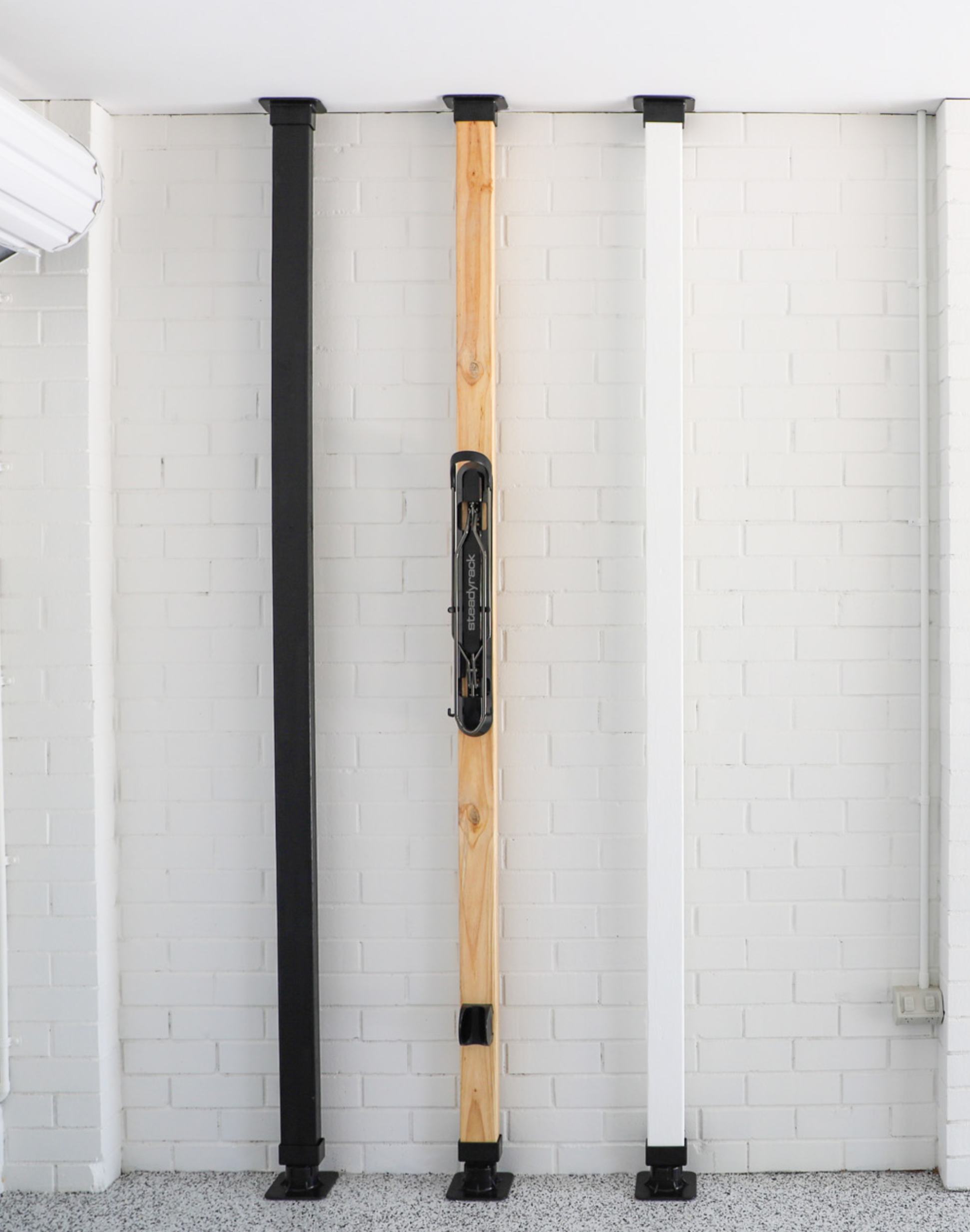 A white, raw and black wooden pole with black extendable bases on the top and bottom, on a white brick background. 